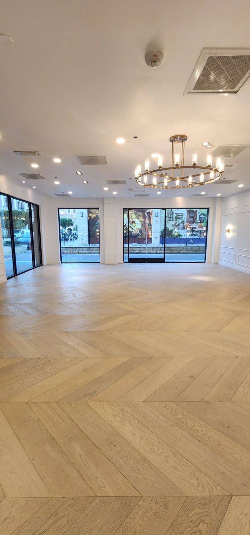 Empty modern interior with herringbone wood flooring and a decorative chandelier. Large windows.