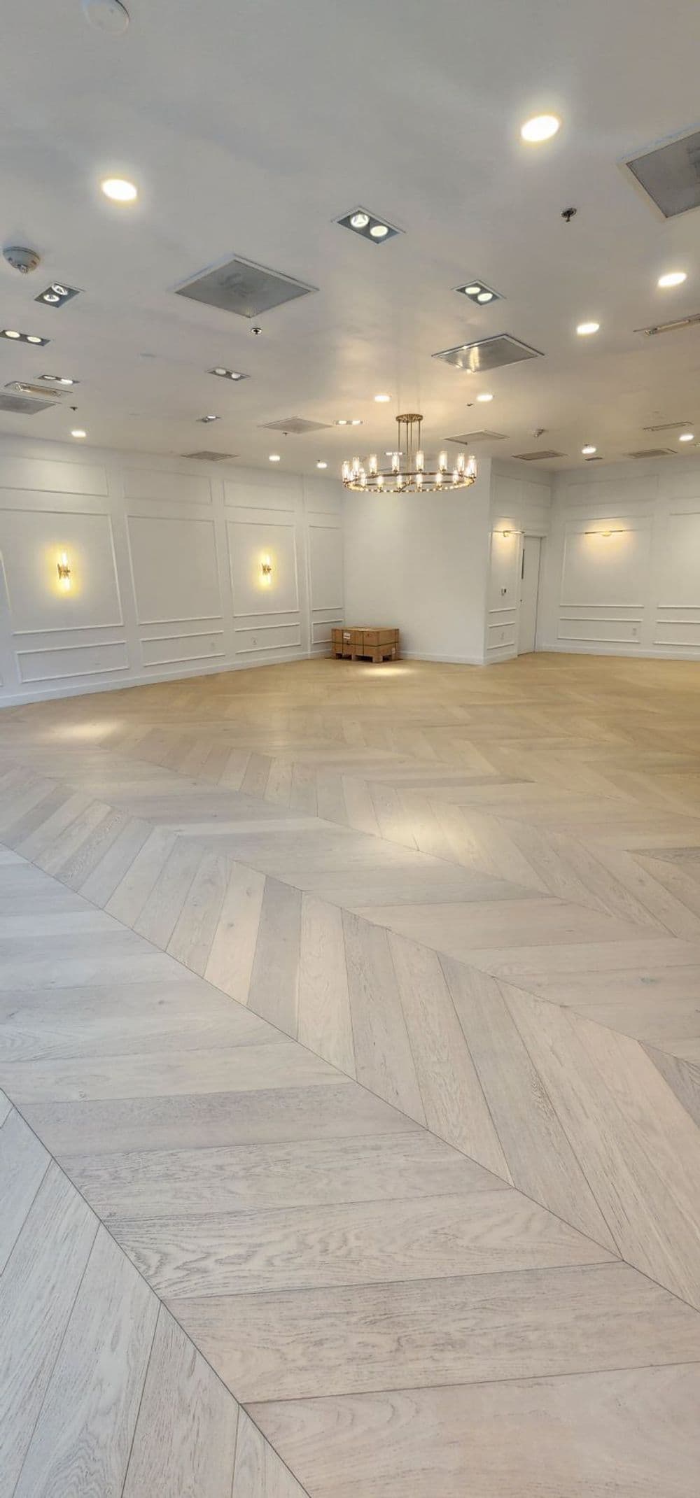 Spacious, well-lit empty room with herringbone wood floor and chandelier.